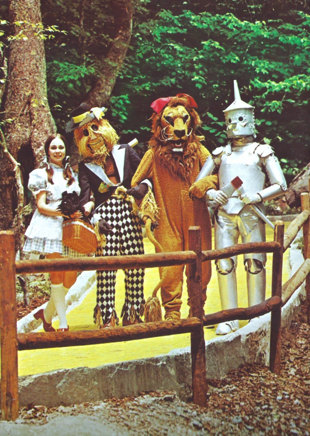 Vintage 1970s photograph of the Wizard of Oz cast — Dorothy, Scarecrow, Cowardly Lion, and Tin Man — posing together by a wooden fence inside Land of Oz Theme Park